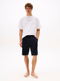 T-shirt bianco in cotone con logo "TOMMY HILFIGER", abbinato a pantaloni corti blu navy con vita elasticizzata e tasche laterali. Modello scalzo.