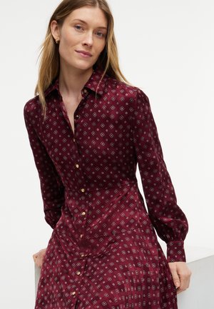 Femme à la peau claire et aux cheveux blonds raides portant une robe bordeaux à manches longues avec un petit motif floral et des boutons dorés.