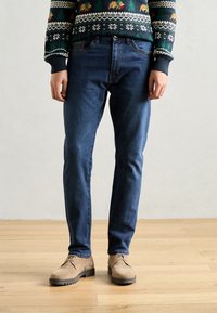 Donkerblauwe slim fit jeans, gemaakt van denim, gecombineerd met beigekleurige suède schoenen. De outfit heeft een gebreide trui met patroon aan de bovenkant.