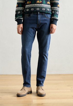 Dark blue jeans with a slim fit, made of denim, paired with beige suede shoes. The outfit features a patterned knitted sweater on top.