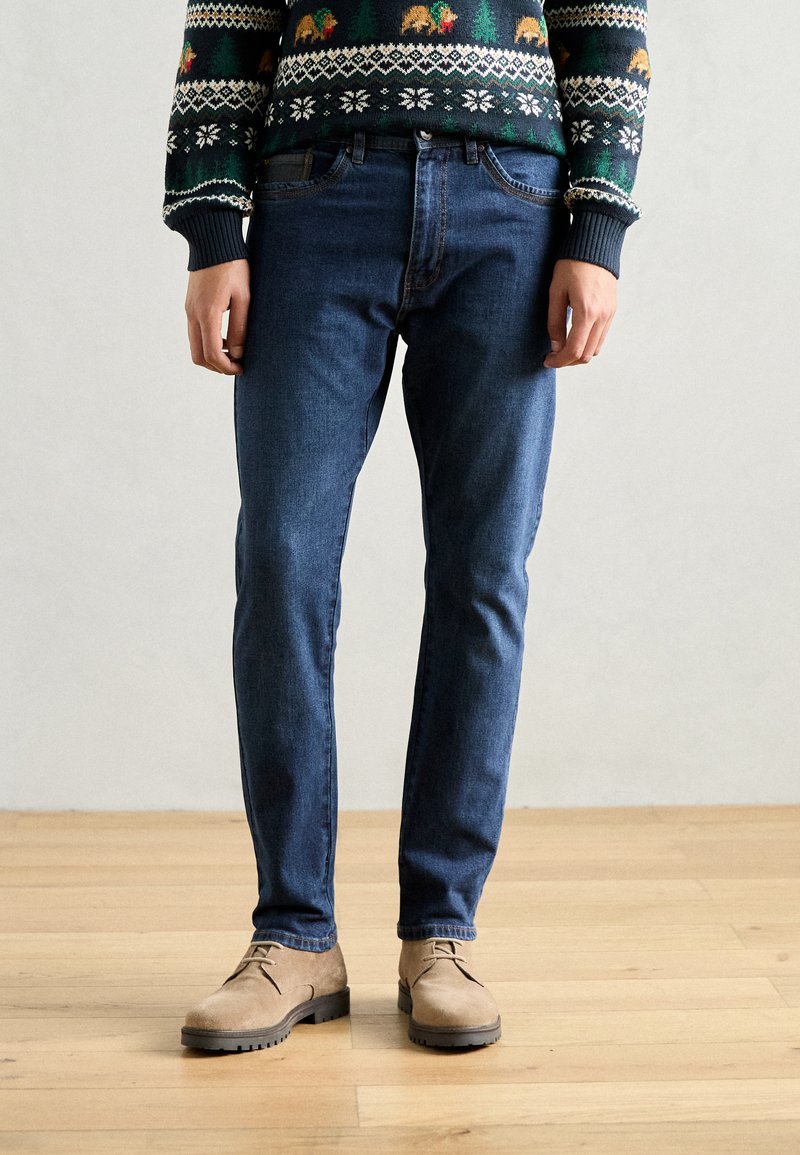 Donkerblauwe slim fit jeans, gemaakt van denim, gecombineerd met beigekleurige suède schoenen. De outfit heeft een gebreide trui met patroon aan de bovenkant.