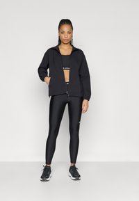 Μαύρο μπουφάν με φερμουάρ, κομμένο μαύρο αθλητικό σουτιέν και γυαλιστερά μαύρα κολάν. Το μοντέλο φοράει μαύρα αθλητικά παπούτσια. Λείο ύφασμα και εφαρμοστή σχεδίαση.