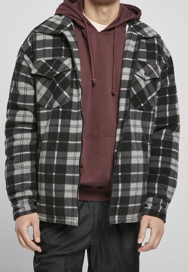 Veste en polaire à carreaux noirs et gris avec deux poches poitrine, ouverte sur un sweat à capuche bordeaux. Texture douce, design décontracté.