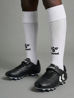 Piernas con calcetas blancas hasta la rodilla de hummel y botas negras de fútbol hummel con tacos sobre un fondo de suelo gris.