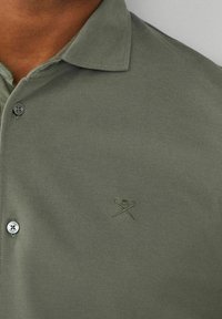 Polo shirt vert olive en tissu texturé, avec un col, une patte de boutonnage à deux boutons et un logo brodé ton sur ton sur la poitrine.
