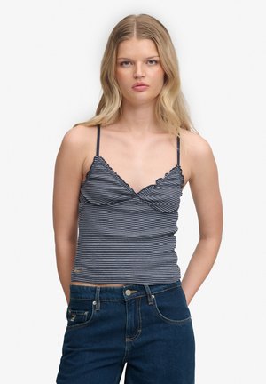 Jeune femme aux cheveux blonds portant un top à fines bretelles bleu marine et blanc à rayures et un jean bleu foncé, debout les mains dans les poches.