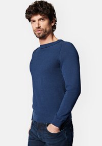 Niebieski sweter z dzianiny o prążkowanej fakturze, z długimi rękawami i okrągłym dekoltem, z subtelnym logo na klatce piersiowej. Noszony z ciemnymi dżinsami.