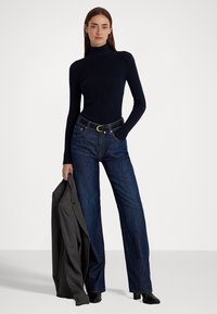 Sweater de gola alta canelado azul marinho, jeans de cintura alta, cinto preto, blazer cinza a pender pelo braço e botins.