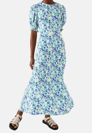 Robe imprimée florale en bleu clair, vert et blanc, avec des manches courtes bouffantes et une silhouette fluide, associée à des sandales blanches épaisses.