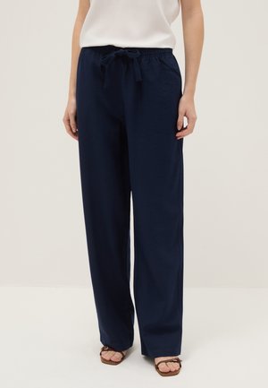 Vrouw draagt marineblauwe broek met wijde pijpen en trekkoord in de taille, gecombineerd met bruine sandalen met open teen, staand tegen een effen achtergrond.