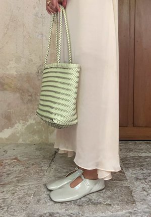 Persoană purtând o rochie lungă crem, ținând o geantă țesută și purtând pantofi Mary Jane lucioși de culoare deschisă pe podea de piatră.