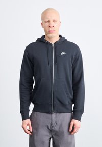 Nike zipzáras kapucnis pulóver sötétszürke színben, puha anyagból készült, elülső zsebbel, húzózsinórral ellátott kapucnival és fehér logóval a bal mellen.