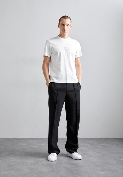 Camisa de algodón blanca con un gráfico simple de montañas, combinada con pantalones negros con plisado y zapatillas deportivas blancas, sobre un fondo gris claro.
