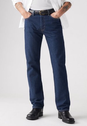 Personne portant un jean bleu foncé à jambes droites avec une ceinture en cuir noire et des bottines noires, mains dans les poches, chemise blanche partiellement visible.