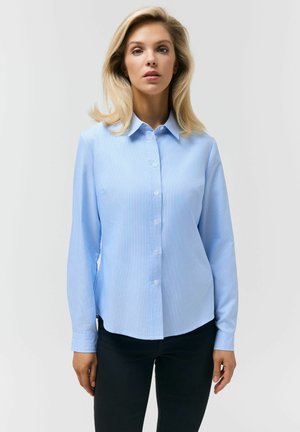 Blondynka w jasnoniebieskiej koszuli na guziki i czarnych spodniach stoi na tle jednolitego białego tła, zwrócona przodem.