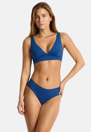 Kobieta ubrana w niebieskie bikini z głębokim dekoltem w kształcie litery V i pasujące dołu, stojąca na jednolitym białym tle.