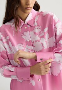 Roze bloes met bloemenpatroon, voorzien van een kraag, lange mouwen en knopen. De stof heeft een gladde textuur met een glanzende afwerking.