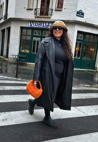Svart läder trench coat, svart figursydd topp, svarta leggings, kraftiga svarta stövlar och en orange plädväska. Urban miljö, blöt asfalt.