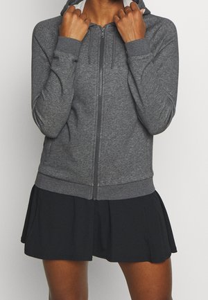 Persona que lleva una sudadera gris con cremallera y cordones, y una falda negra plisada, con las manos sujetando el cuello de la sudadera.