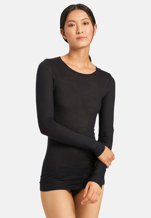 Donna con capelli scuri legati in una coda bassa, che indossa una maglietta nera aderente a maniche lunghe, in piedi con le braccia incrociate davanti a uno sfondo semplice.