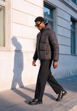 Homme portant une doudoune noire, un pantalon noir et des bottes marche sur le trottoir de la ville à côté d’un bâtiment avec de grandes fenêtres en plein jour.