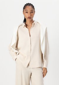 Beige Knopfhemd aus einem weichen, strukturierten Stoff mit einer lockeren Passform. Ein weißer Pullover ist über die Schultern drapiert.