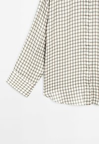 Chemise à manches longues avec un motif à carreaux beige et marron sur tissu blanc, manche retroussée visible sur le côté gauche.