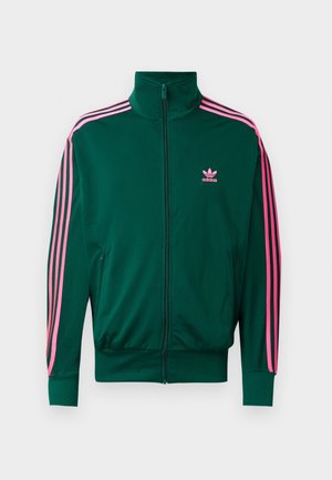 Zielona kurtka Adidas na zamek błyskawiczny, z różowymi paskami na rękawach i różowym logo Adidas po lewej stronie klatki piersiowej, pokazana na białym tle.