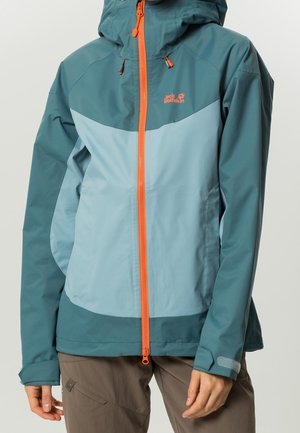 Veste imperméable bicolore bleue avec fermeture éclair orange et capuche ajustable portée avec un pantalon marron, visible des épaules aux hanches.