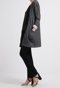 Grå, texturerad cardigan med oversize passform och två framfickor, lagd över en svart och vit randig topp och svarta byxor, parat med guld sandaler.