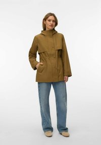 Femme portant une veste de pluie marron à capuche avec des boutons-pression, un jean bleu et des chaussures beige, debout devant un fond blanc.