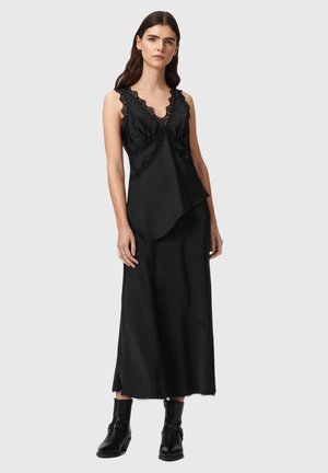 Kvinde iført sort ærmeløs top med blonder og lang sort nederdel, kombineret med sorte ankelstøvler, stående mod en ensfarvet baggrund.