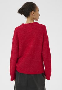 Punane kootud kampsun lõdva lõikega, langevate õlgade ja soonikkoes äärega. Materjal tundub pehme, tagasihoidlikult tekstuuriga viimistlusega.