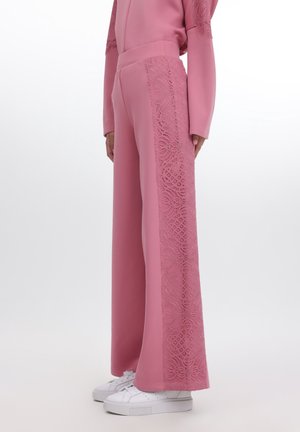 Kobieta ubrana w różowe spodnie z szerokimi nogawkami z koronkowymi panelami po bokach oraz pasujący długi rękaw, w połączeniu z białymi trampkami, stojąca bokiem.