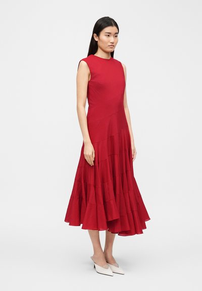 Γυναίκα που στέκεται φορώντας ένα αμάνικο, κοκκινωπό φόρεμα μήκους μέχρι το γόνατο με πολύπλοκη φούστα και λευκά τακούνια με μυτερή μύτη σε απλό φόντο.