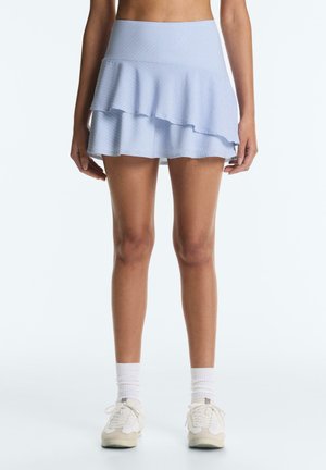 Kobieta ubrana w jasnoniebieską warstwową mini spódniczkę, białe skarpetki i białe sportowe buty, stoi na jednolitym tle.