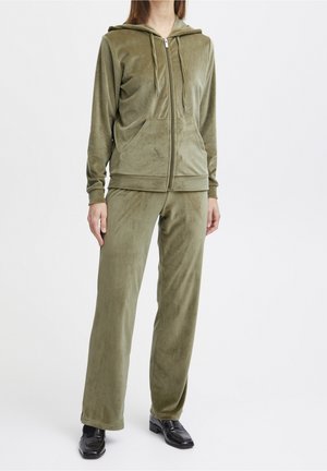 Osoba ubrana w oliwkowozieloną welurową bluzę z zamkiem oraz pasujące szerokie spodnie nogawki, nosząca czarne loafersy, stojąca na jednolitym tle.