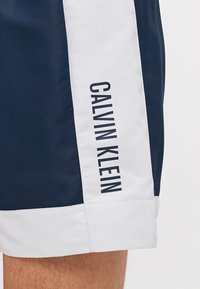 Mornarsko modri in beli kopalni šorti z logotipom, natisnjenim vertikalno na strani. Narejeni so iz gladkega materiala s čistim, športnim dizajnom.