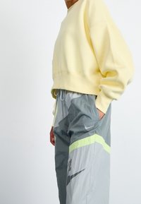 En person iklädd en ljusgul sweatshirt och grå Nike träningbyxor med neongröna detaljer, med händerna i fickorna, mot en enkel vit bakgrund.