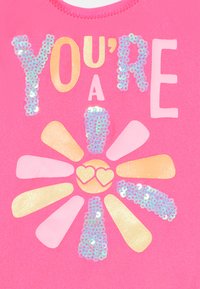 Fond rose vif avec des lettres en sequins métalliques dorés et multicolores indiquant "TU ES UN" et un motif de fleur avec des accents en forme de cœur.