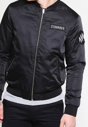 Mand iført en sort bomberjakke med lynlås, med et mærke med teksten "C109R9E0" på brystet og et rundt mærke på ærmet.