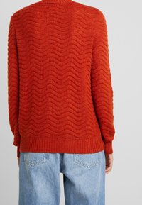 Texturovaný svetr v cihlově červené barvě s vlnovým vzorem, žebrovanými manžetami a lemem. V kombinaci s světle modrými volnými džíny, které ukazují uvolněný střih.