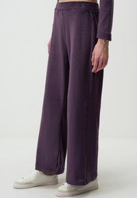Pantalons larges violets en tissu doux et texturé, avec une taille haute et des détails de couture subtils. Associés à des baskets blanches.