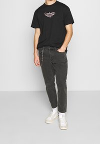 Czarna bawełniana koszulka z białym logo „Carhartt”, zestawiona z ciemnoszarymi dżinsami z detalem łańcucha oraz białymi trampkami w minimalistycznym otoczeniu.