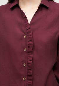 Μπορντό πουκάμισο με κουμπιά, διαθέτει φρακισταρίδα, τέσσερα στρογγυλά κουμπιά και κλασικό γιακά. Λείος, υφασμάτινος ιστός.