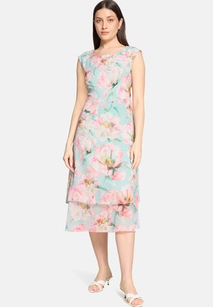 Vrouw draagt een mouwloze, lichtblauwe midi-jurk met een roze en witte bloemenprint, gecombineerd met witte open-toe sandalen met hak.