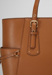 Sac à main en cuir marron texturé avec une bandoulière réglable dotée d'une boucle et de détails métalliques dorés, sur un fond uni.