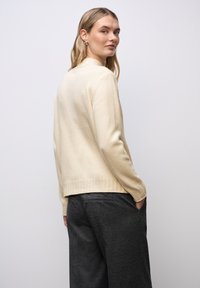 Pull en beige tricoté avec un ourlet et des poignets côtelés, présentant une coupe décontractée et des manches longues, porté sur un pantalon gris foncé.