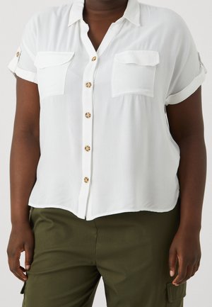 Mujer con una camisa blanca de manga corta con botones y dos bolsillos en el pecho y pantalones de color verde oliva.