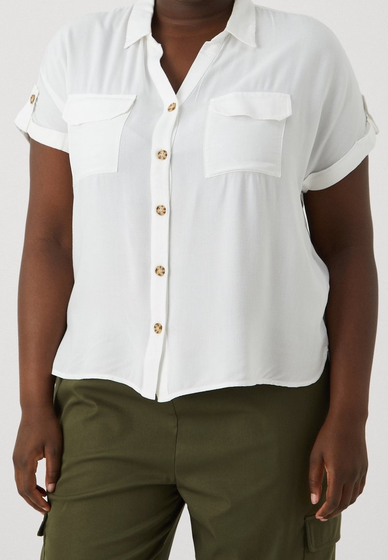 Femme portant une chemise blanche à manches courtes avec deux poches poitrine et un pantalon vert olive.